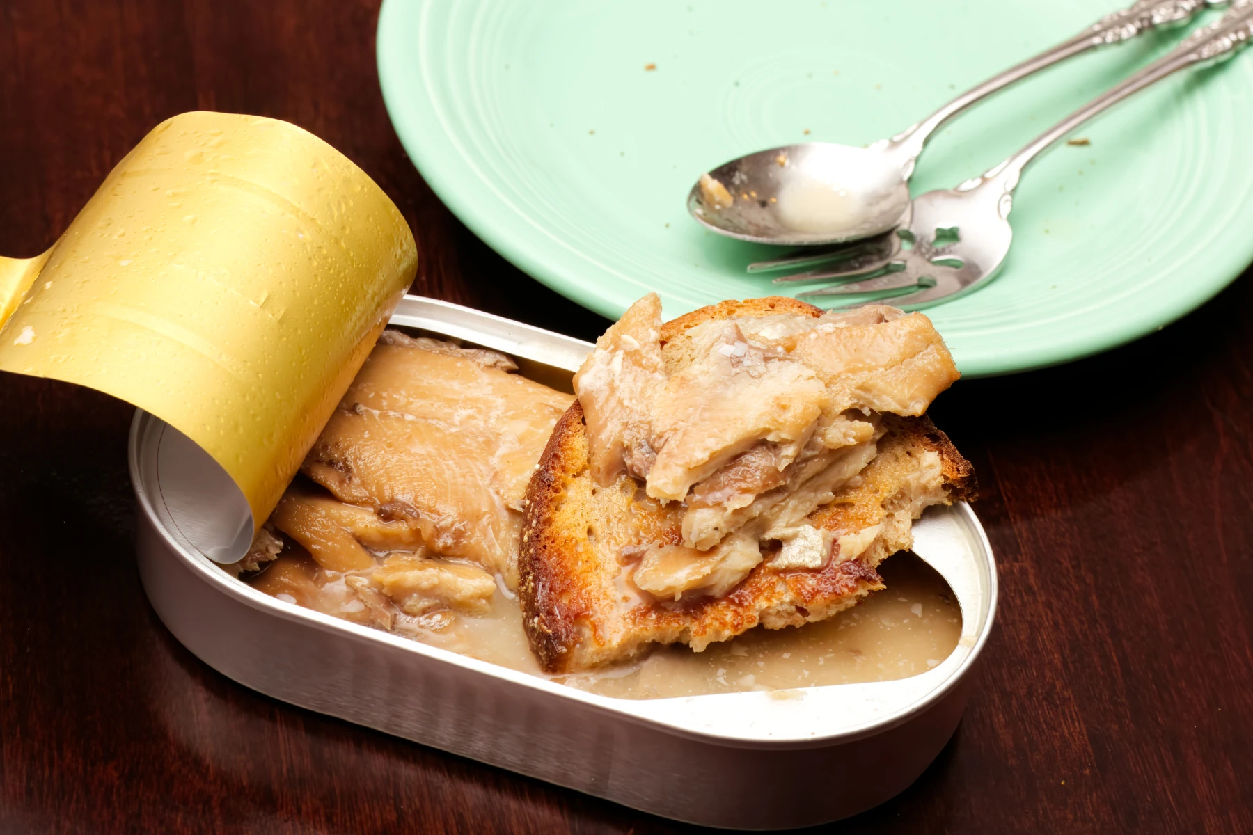 Half eaten bread with sardine morsels, empty plate and near empty tin.