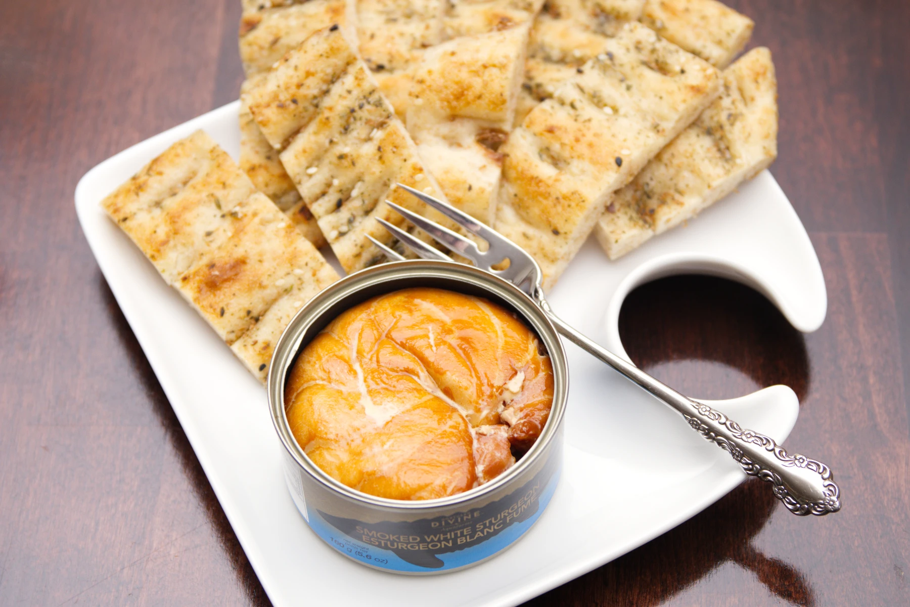 Opened tin of Northern Divine Smoked White Sturgeon on a plate with garlic flatbread