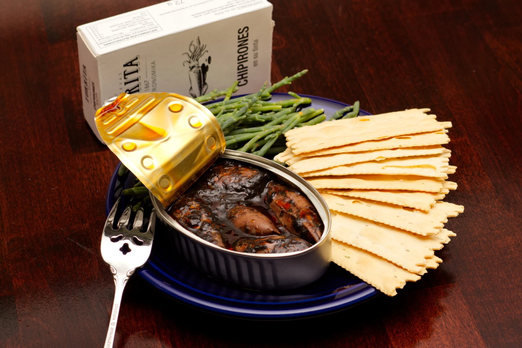 Opened can of squid on a plate with crackers and some sea asparagus.